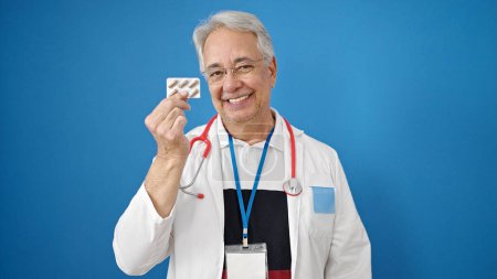 Photo for Middle age man with grey hair doctor holding pills over isolated blue background - Royalty Free Image