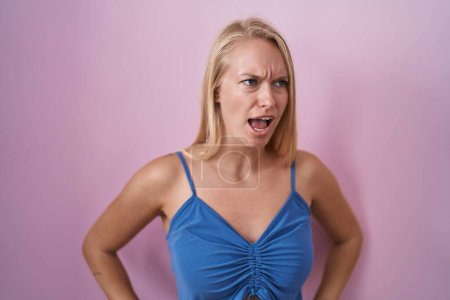 Foto de Young caucasian woman standing over pink background angry and mad screaming frustrated and furious, shouting with anger. rage and aggressive concept. - Imagen libre de derechos