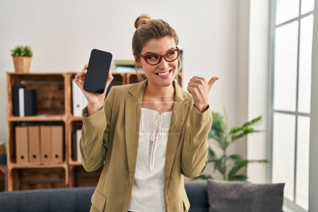 Téléchargez les photos : Jeune femme travaillant au bureau de consultation tenant smartphone pointant le pouce vers le côté souriant heureux avec la bouche ouverte - en image libre de droit