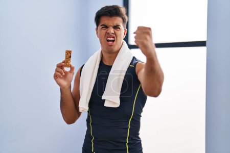 Téléchargez les photos : Hispanique homme manger barre de protéines comme collation énergie saine ennuyé et frustré criant avec colère, hurlant fou de colère et la main levée - en image libre de droit