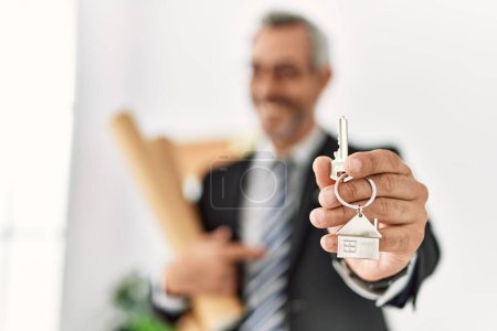 Foto de Arquitecto hombre de pelo gris de mediana edad sosteniendo planos y llave de casa nueva en la oficina - Imagen libre de derechos