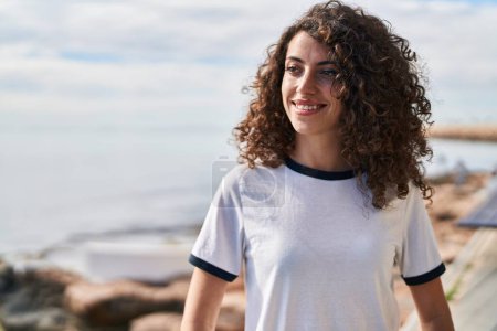 Foto de Mujer hispana joven sonriendo confiada mirando a un lado a la orilla del mar - Imagen libre de derechos