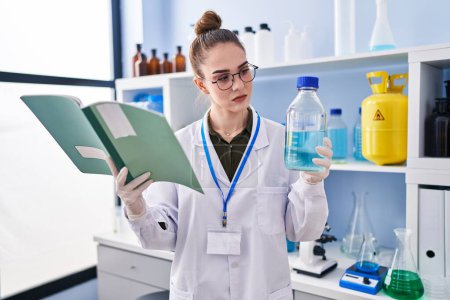 Téléchargez les photos : Young woman scientist reading book holding bottle at laboratory - en image libre de droit