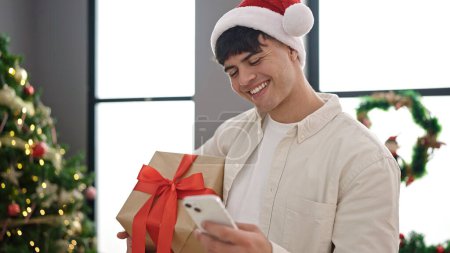 Foto de Joven hombre hispano usando smartphone sosteniendo regalo de Navidad en casa - Imagen libre de derechos