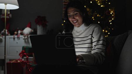 Téléchargez les photos : Jeune belle femme hispanique en utilisant un ordinateur portable célébrant Noël à la maison - en image libre de droit