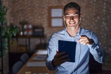 Téléchargez les photos : Bel homme hispanique travaillant au bureau la nuit vous montrant du doigt et la caméra, souriant positif et joyeux - en image libre de droit