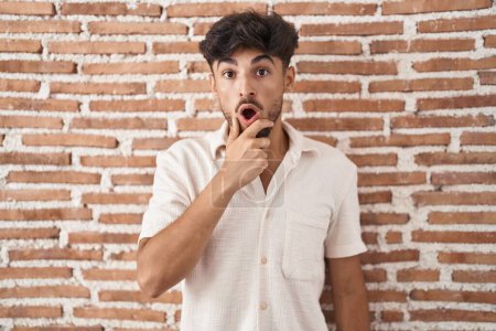 Photo for Arab man with beard standing over bricks wall background looking fascinated with disbelief, surprise and amazed expression with hands on chin - Royalty Free Image