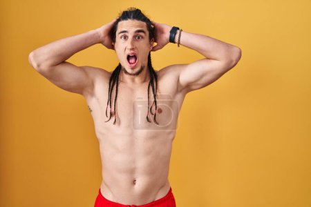 Photo for Hispanic man with long hair standing shirtless over yellow background crazy and scared with hands on head, afraid and surprised of shock with open mouth - Royalty Free Image