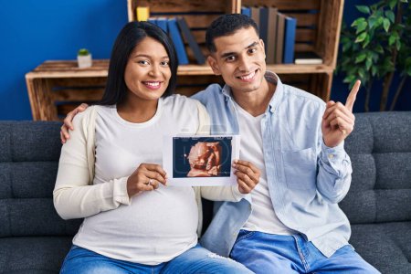 Photo for Young hispanic couple expecting a baby sitting on the sofa showing baby ultrasound smiling happy pointing with hand and finger to the side - Royalty Free Image