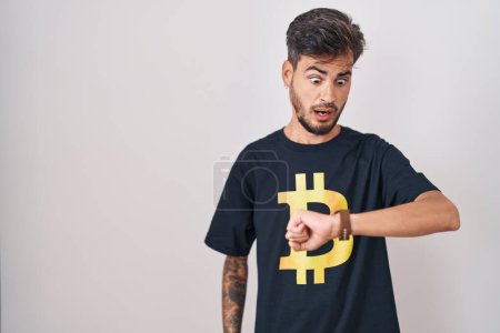 Foto de Joven hombre hispano con tatuajes con camiseta Bitcoin mirando el tiempo del reloj preocupado, con miedo de llegar tarde - Imagen libre de derechos