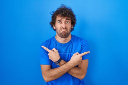 Téléchargez les photos : Jeune homme hispanique debout sur fond bleu pointant des deux côtés avec les doigts, direction différente en désaccord - en image libre de droit