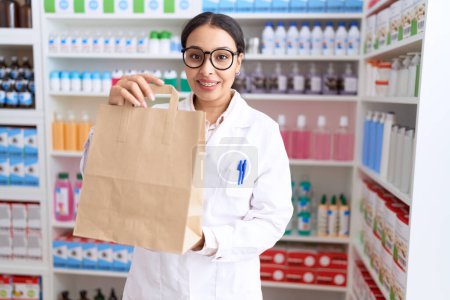Téléchargez les photos : Jeune pharmacienne arabe souriante confiante tenant son sac à provisions à la pharmacie - en image libre de droit