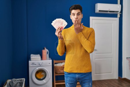 Photo for Young hispanic man at laundry room holding shekels covering mouth with hand, shocked and afraid for mistake. surprised expression - Royalty Free Image
