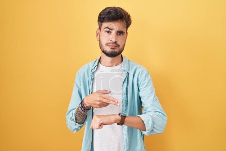 Téléchargez les photos : Jeune homme hispanique avec des tatouages debout sur fond jaune à la hâte pointant vers regarder le temps, l'impatience, bouleversé et en colère pour retard de délai - en image libre de droit
