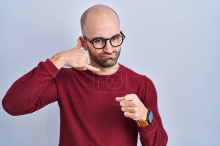 Photo for Young bald man with beard standing over white background wearing glasses smiling doing talking on the telephone gesture and pointing to you. call me. - Royalty Free Image