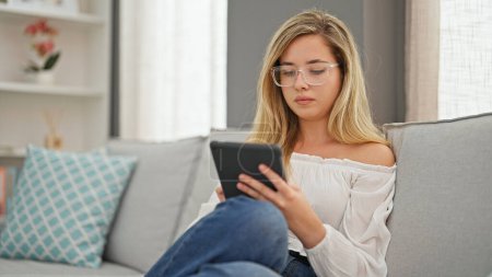 Téléchargez les photos : Jeune femme blonde portant des lunettes à l'aide de touchpad à la maison - en image libre de droit
