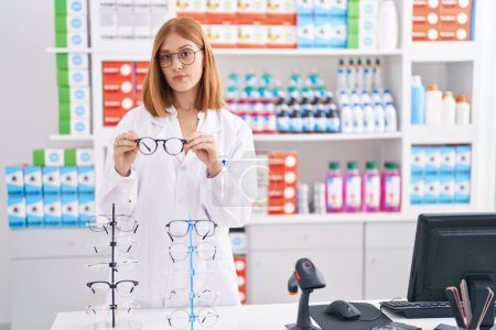 Photo for Young redhead woman working at pharmacy drugstore holding glasses relaxed with serious expression on face. simple and natural looking at the camera. - Royalty Free Image