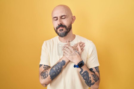 Photo for Hispanic man with tattoos standing over yellow background smiling with hands on chest with closed eyes and grateful gesture on face. health concept. - Royalty Free Image