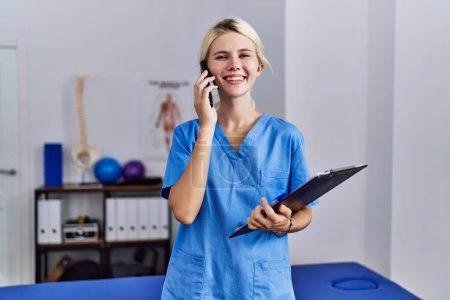 Téléchargez les photos : Jeune femme blonde pysiothérapeute parlant sur smartphone tenant presse-papiers à la clinique de réadaptation - en image libre de droit