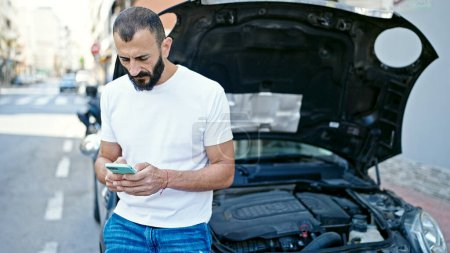 Foto de Hombre hispano joven mensajes de texto con seguro por teléfono inteligente para la rotura del coche en la calle - Imagen libre de derechos