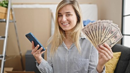 Foto de Mujer rubia joven usando smartphone sosteniendo dólares en nuevo hogar - Imagen libre de derechos