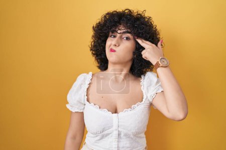 Photo for Young brunette woman with curly hair standing over yellow background shooting and killing oneself pointing hand and fingers to head like gun, suicide gesture. - Royalty Free Image