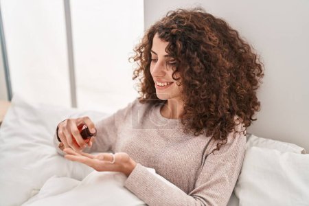 Téléchargez les photos : Jeune femme hispanique versant de la lotion sur la main assise sur le lit dans la chambre - en image libre de droit