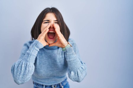 Photo for Young hispanic woman standing over blue background shouting angry out loud with hands over mouth - Royalty Free Image