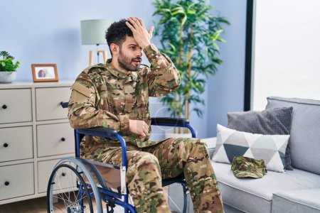 Photo for Arab man wearing camouflage army uniform sitting on wheelchair surprised with hand on head for mistake, remember error. forgot, bad memory concept. - Royalty Free Image