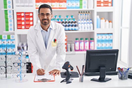 Téléchargez les photos : Jeune homme hispanique pharmacien souriant confiant écrit sur le document à la pharmacie - en image libre de droit