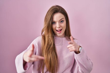 Foto de Young caucasian woman standing over pink background pointing fingers to camera with happy and funny face. good energy and vibes. - Imagen libre de derechos
