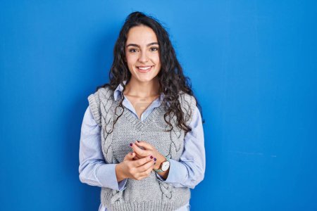 Téléchargez les photos : Jeune femme brune debout sur fond bleu avec les mains jointes et les doigts croisés souriant détendu et joyeux. succès et optimisme - en image libre de droit