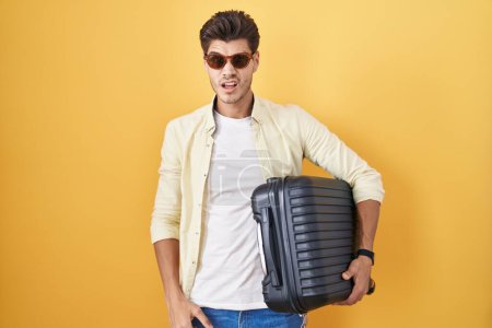 Téléchargez les photos : Jeune homme hispanique tenant une valise allant en vacances d'été dans le visage de choc, l'air sceptique et sarcastique, surpris avec la bouche ouverte - en image libre de droit