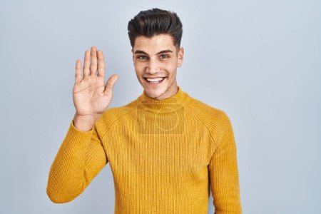 Téléchargez les photos : Jeune homme hispanique debout sur fond bleu renonçant à dire bonjour heureux et souriant, geste de bienvenue amical - en image libre de droit