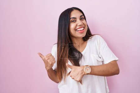 Téléchargez les photos : Jeune femme arabe debout sur fond rose pointant vers le dos derrière avec la main et les pouces vers le haut, souriant confiant - en image libre de droit