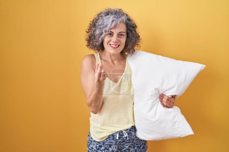 Téléchargez les photos : Femme d'âge moyen aux cheveux gris portant un pyjama étreignant un oreiller faisant un geste d'argent avec les mains, demandant un paiement de salaire, entreprise millionnaire - en image libre de droit