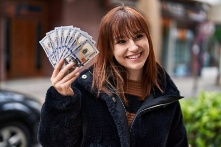 Foto de Joven mujer hermosa sosteniendo billetes de dólares con aspecto positivo y feliz de pie y sonriendo con una sonrisa confiada mostrando los dientes - Imagen libre de derechos