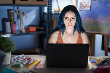 Photo for Young modern girl with blue hair sitting at art studio with laptop at night depressed and worry for distress, crying angry and afraid. sad expression. - Royalty Free Image
