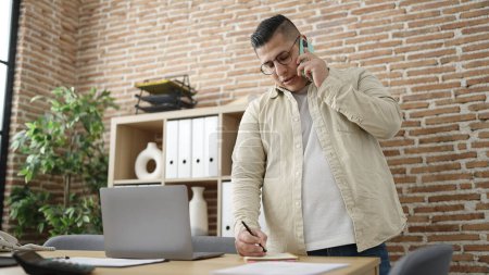 Foto de Joven trabajador de negocios hispano hablando por teléfono inteligente escribiendo en un cuaderno en la oficina - Imagen libre de derechos