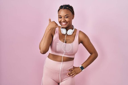 Photo for African american woman with braids wearing sportswear and headphones smiling doing phone gesture with hand and fingers like talking on the telephone. communicating concepts. - Royalty Free Image