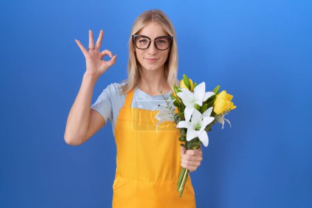Téléchargez les photos : Jeune femme caucasienne portant tablier de fleuriste tenant des fleurs souriant positif faire ok signe avec la main et les doigts. expression réussie. - en image libre de droit