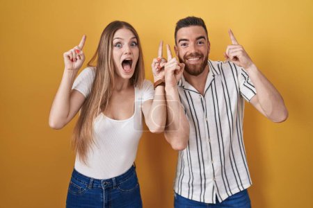 Photo for Young couple standing over yellow background smiling amazed and surprised and pointing up with fingers and raised arms. - Royalty Free Image