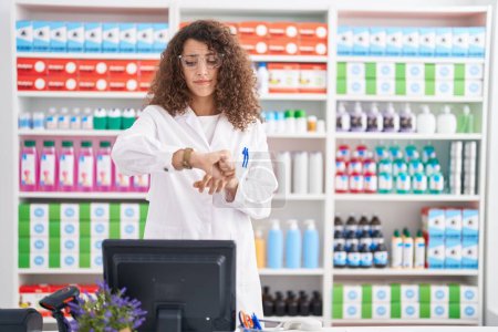 Téléchargez les photos : Femme hispanique aux cheveux bouclés travaillant à la pharmacie pharmacie vérifiant l'heure sur la montre-bracelet, détendue et confiante - en image libre de droit