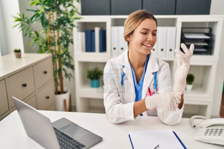 Photo for Young hispanic woman wearing doctor uniform wearing gloves at clinic - Royalty Free Image