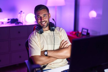 Téléchargez les photos : Jeune homme hispanique banderole souriant confiant assis avec les bras croisés geste à la salle de jeux - en image libre de droit