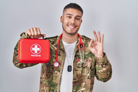 Foto de Joven médico hispano vistiendo uniforme camuflaje del ejército sosteniendo botiquín de primeros auxilios haciendo buen signo con los dedos, sonriente gesto amistoso excelente símbolo - Imagen libre de derechos