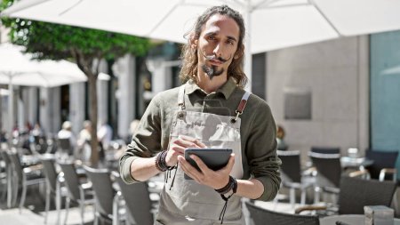 Foto de Joven camarero hispano usando touchpad con cara seria en la terraza de la cafetería - Imagen libre de derechos