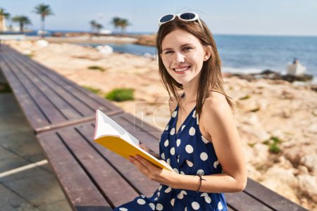 Foto de Joven mujer rubia turista leyendo libro sentado en el banco a la orilla del mar - Imagen libre de derechos