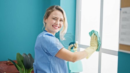 Foto de Mujer rubia joven limpiador profesional ventana de limpieza sonriendo en la oficina - Imagen libre de derechos