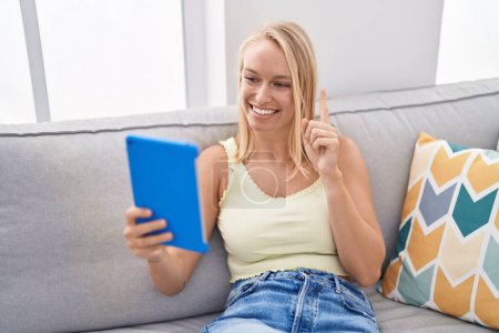 Foto de Joven mujer caucásica usando touchpad sentado en el sofá sonriendo con una idea o pregunta señalando el dedo con la cara feliz, número uno - Imagen libre de derechos